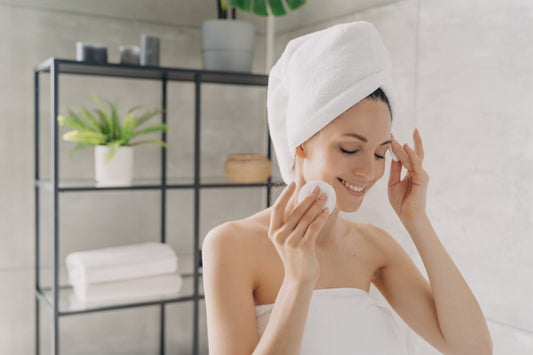 Woman in a white towel and hair wrap smiling while using a reusable facial cleanse pad in a modern bathroom – eco-friendly skincare routine