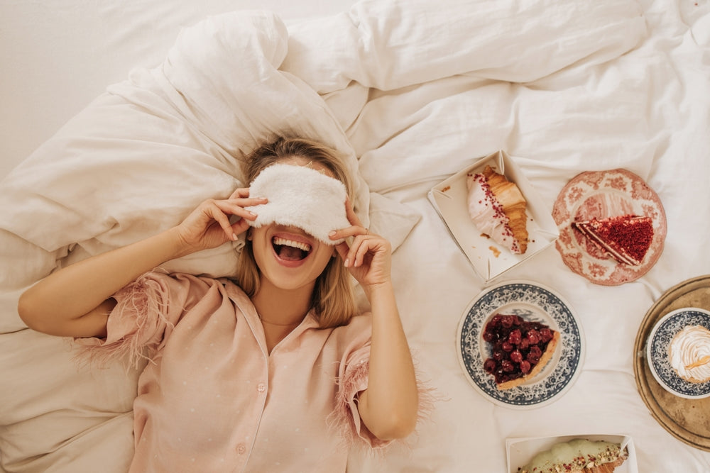 Person relaxing in bed wearing a fluffy white sleep mask, surrounded by plates of cake and dessert, enjoying a cosy self-care moment
