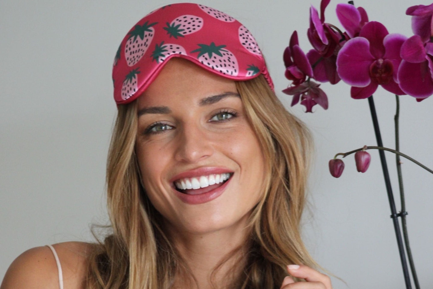 Woman smiling in pink strawberry print cloud satin sleep mask from SMUG Cloud Collection, shown with orchid flowers